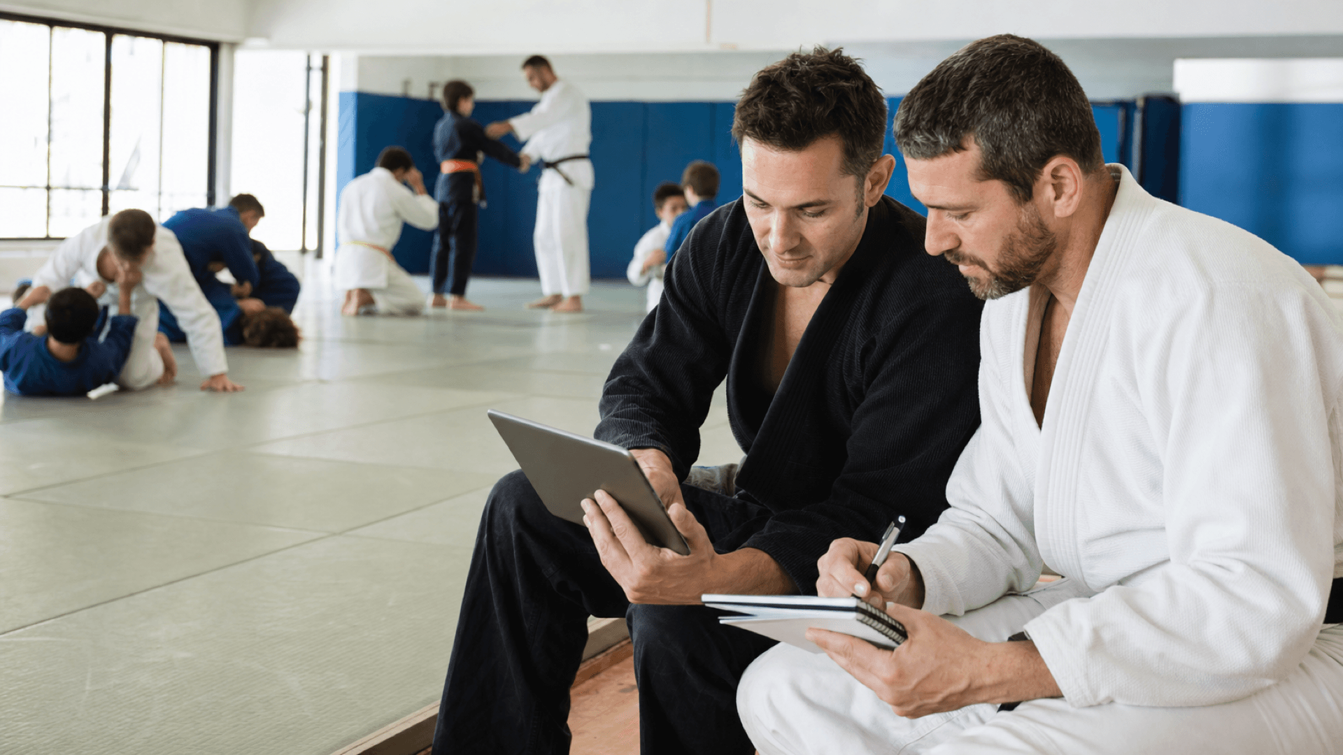 Two instructors plan beside the mat while a group trains in their academy